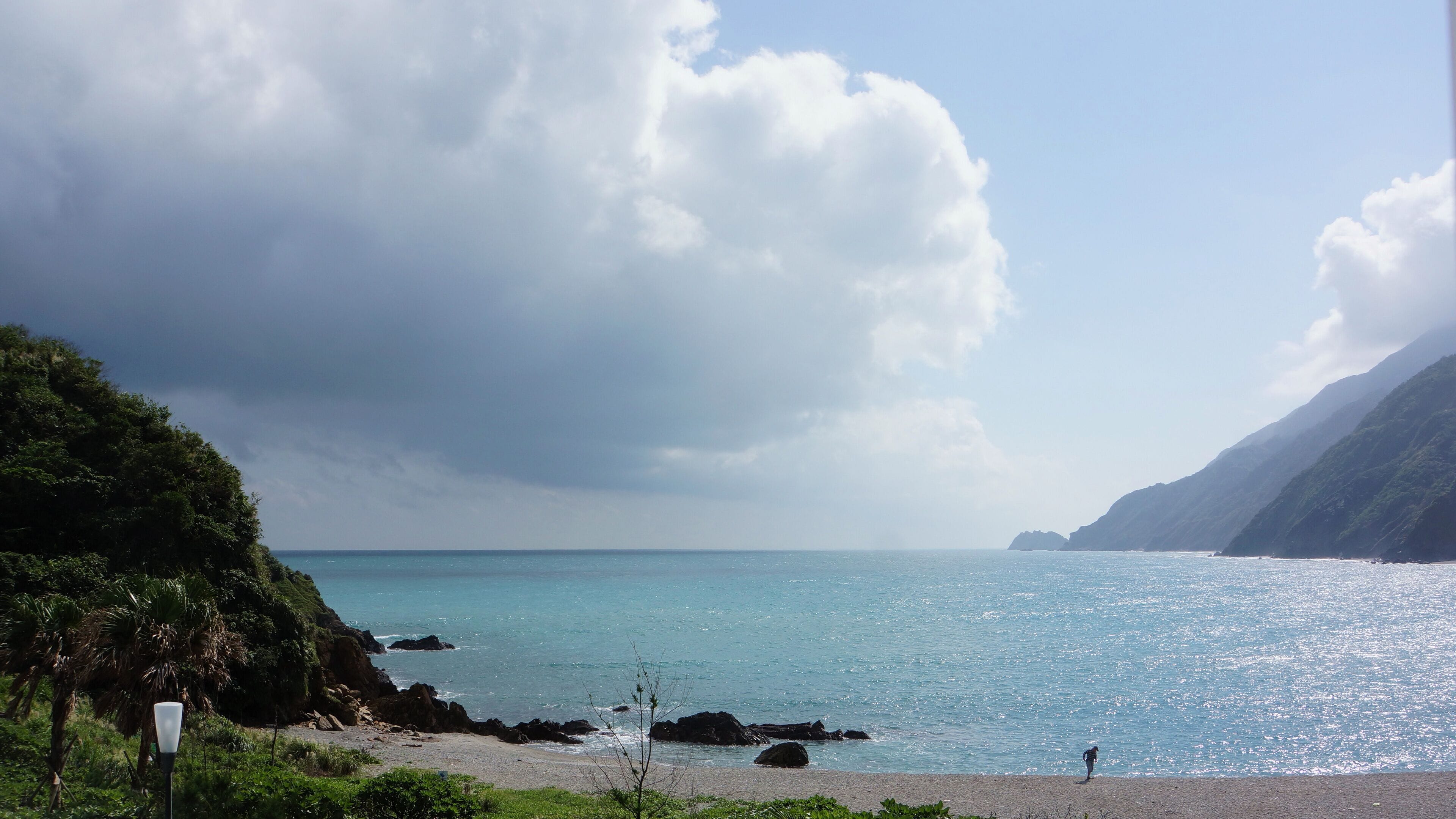 Paisaje costero con playa, costa rocosa y una persona de pie sobre la arena.