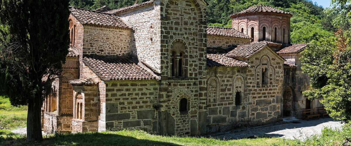 A stone church with a tiled roof, surrounded by lush greenery and a forested hill in the background.
