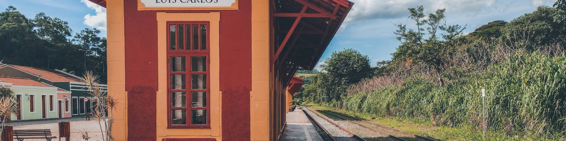 A small, single-story train station named 'LUÍS CARLOS' with a red roof and yellow walls, surrounded by greenery and a clear sky.