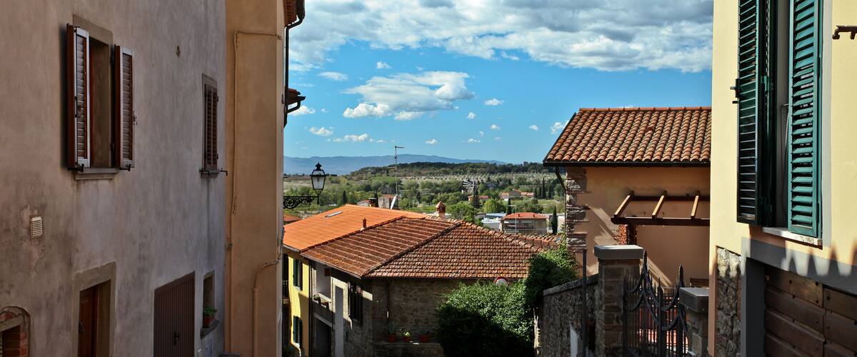 A narrow street in a historic town with terracotta-roofed buildings and a clear blue sky.