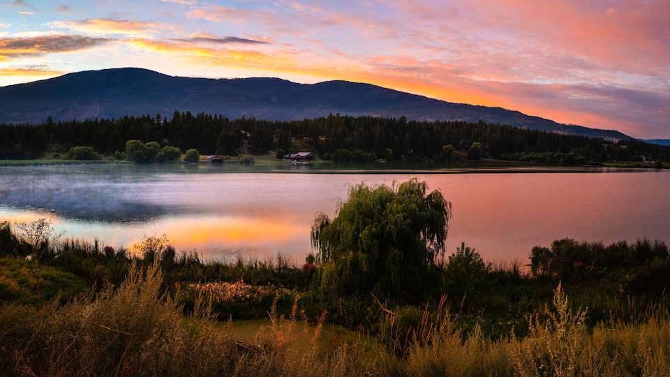 A calm lake with a forested mountain range in the background and a small building near the water's edge.