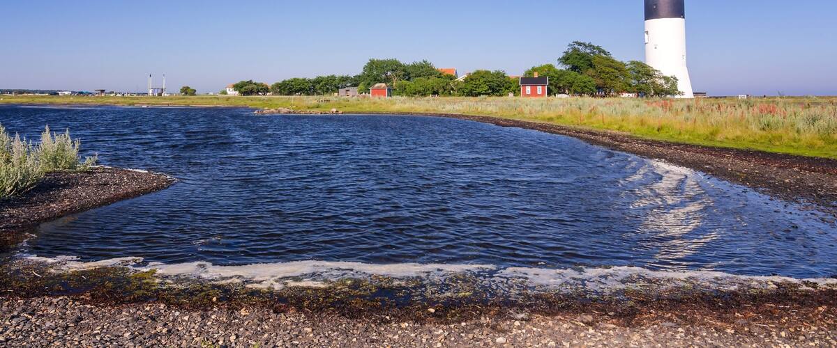 A lighthouse by a calm water body with a rocky shoreline and a red house in the background.