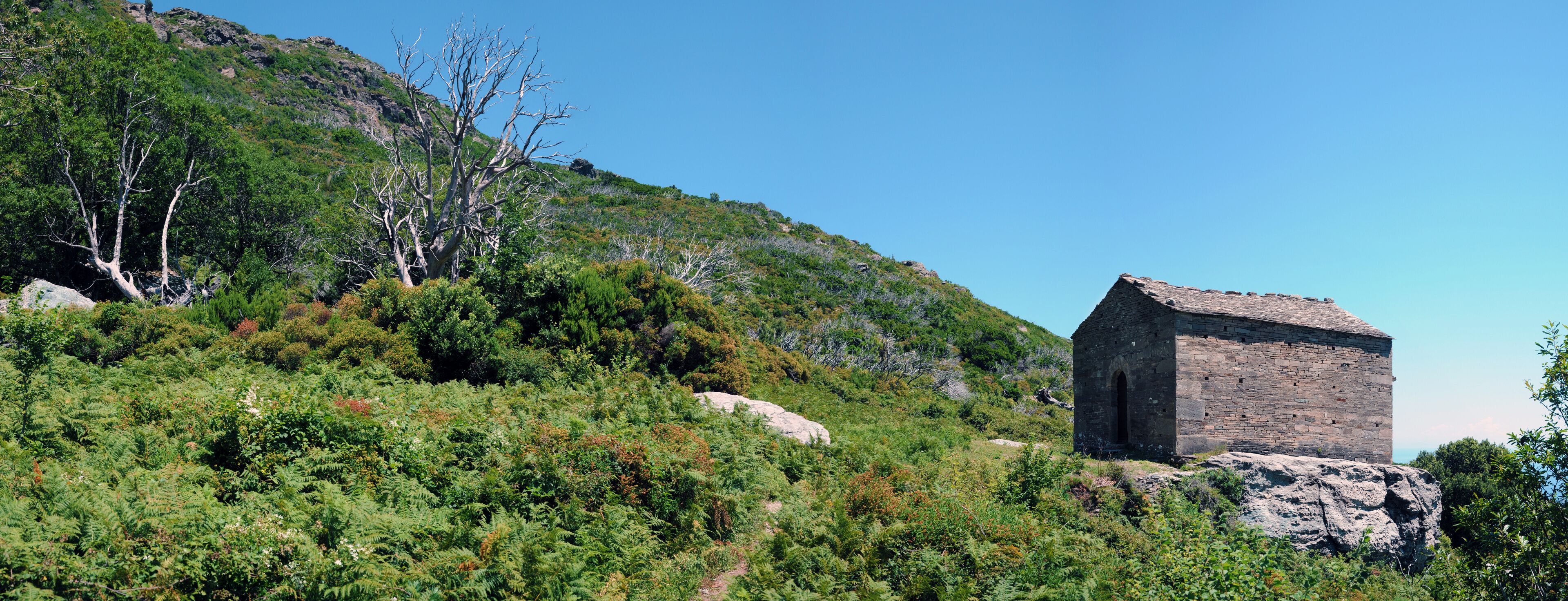 A stone building on a hillside with dense green vegetation and a clear blue sky.