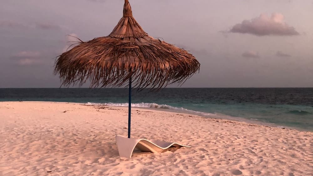 A beach with a thatched umbrella and a white lounge chair.