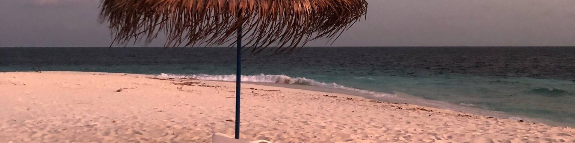 A beach with a thatched umbrella and a white lounge chair.