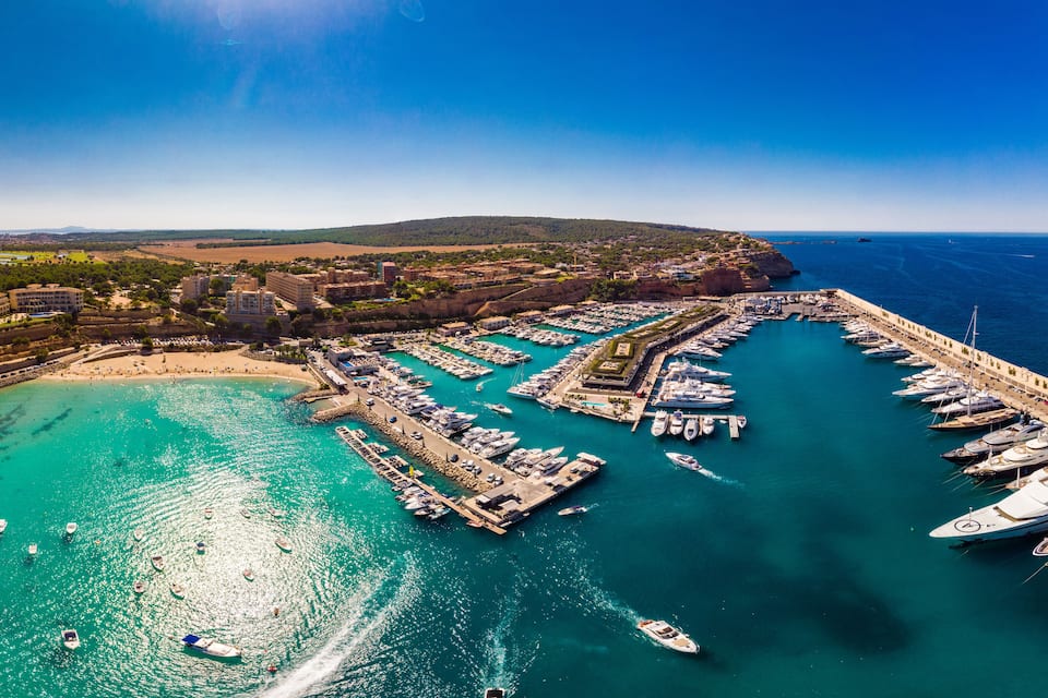 Un port de plaisance situé sur la côte, où de nombreux bateaux sont amarrés, avec une plage de sable et, en arrière-plan, des collines et des bâtiments.