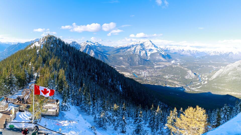 A snow-covered mountain with a Canadian flag, a forested valley, and distant snow-capped peaks.