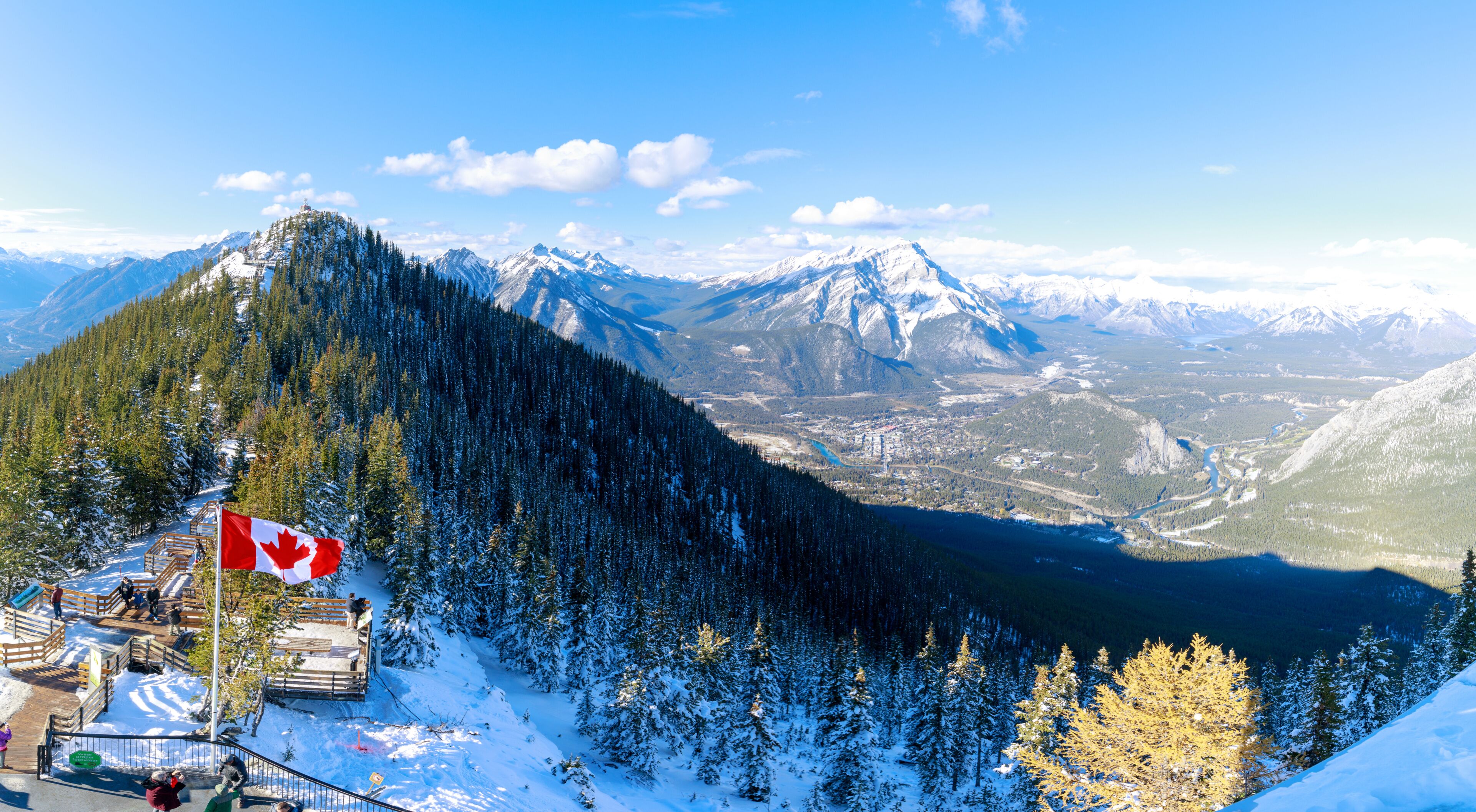 A snow-covered mountain with a Canadian flag, a forested valley, and distant snow-capped peaks.