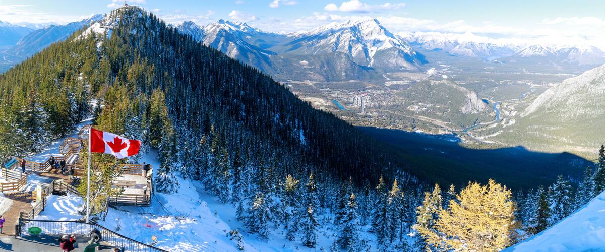 A snow-covered mountain with a Canadian flag, a forested valley, and distant snow-capped peaks.