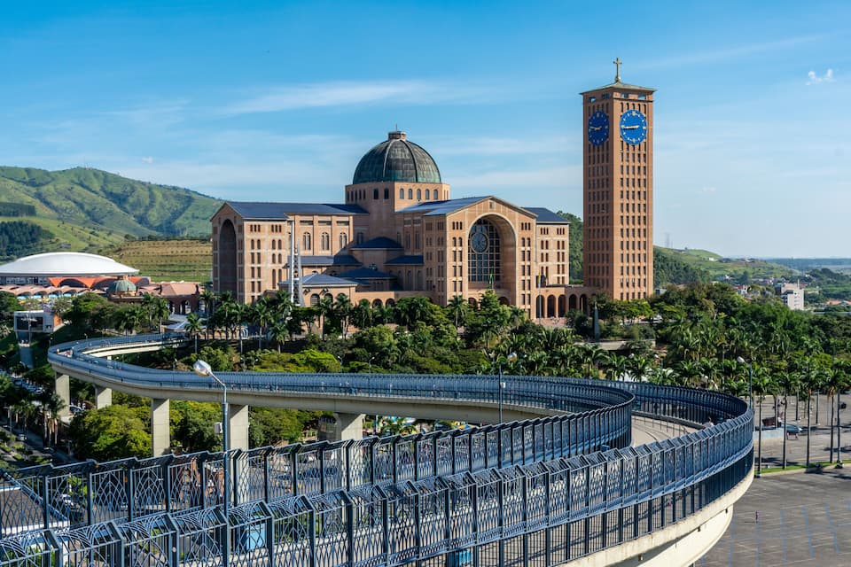 Uma igreja imponente com uma torre do relógio destacada e uma cúpula, rodeada por vegetação exuberante e uma ponte curva.