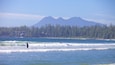 A coastal scene with waves, a surfer, and a forested mountain range in the background.