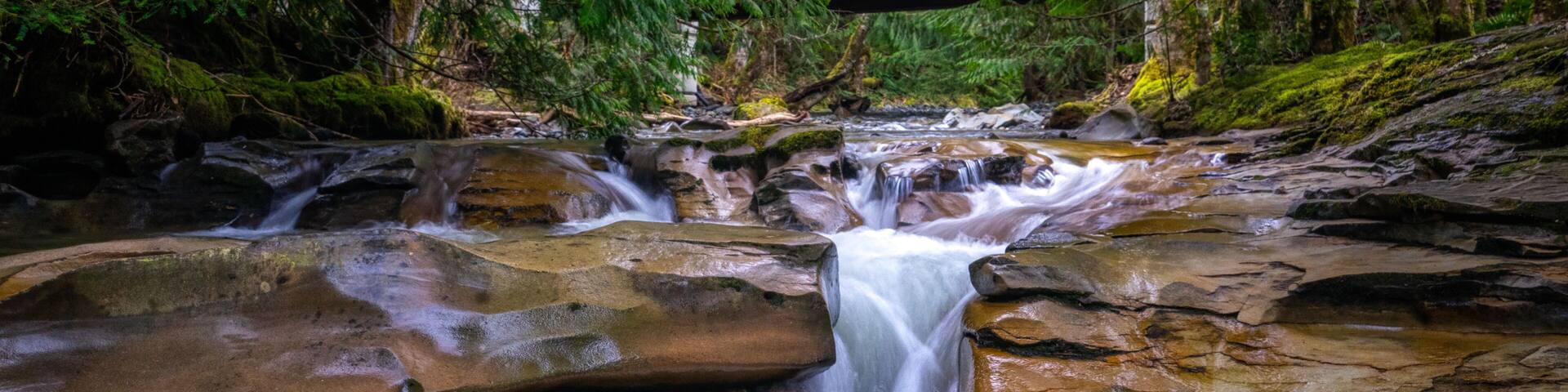 A waterfall cascading over rocks in a forested area with a wooden bridge in the background.
