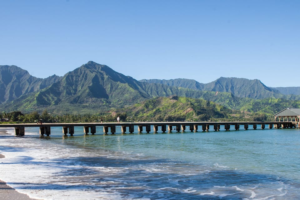 Una spiaggia con onde, un molo che si protende nell'acqua e montagne sullo sfondo.
