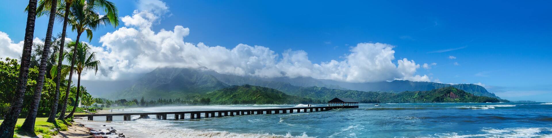A beach with a wooden pier extending into the ocean, palm trees, and a mountain range in the background.