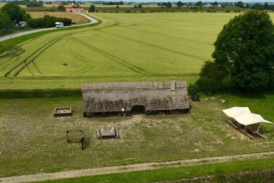 Un paisaje rural con un extenso campo, una construcción pequeña, un río y una casa a lo lejos.