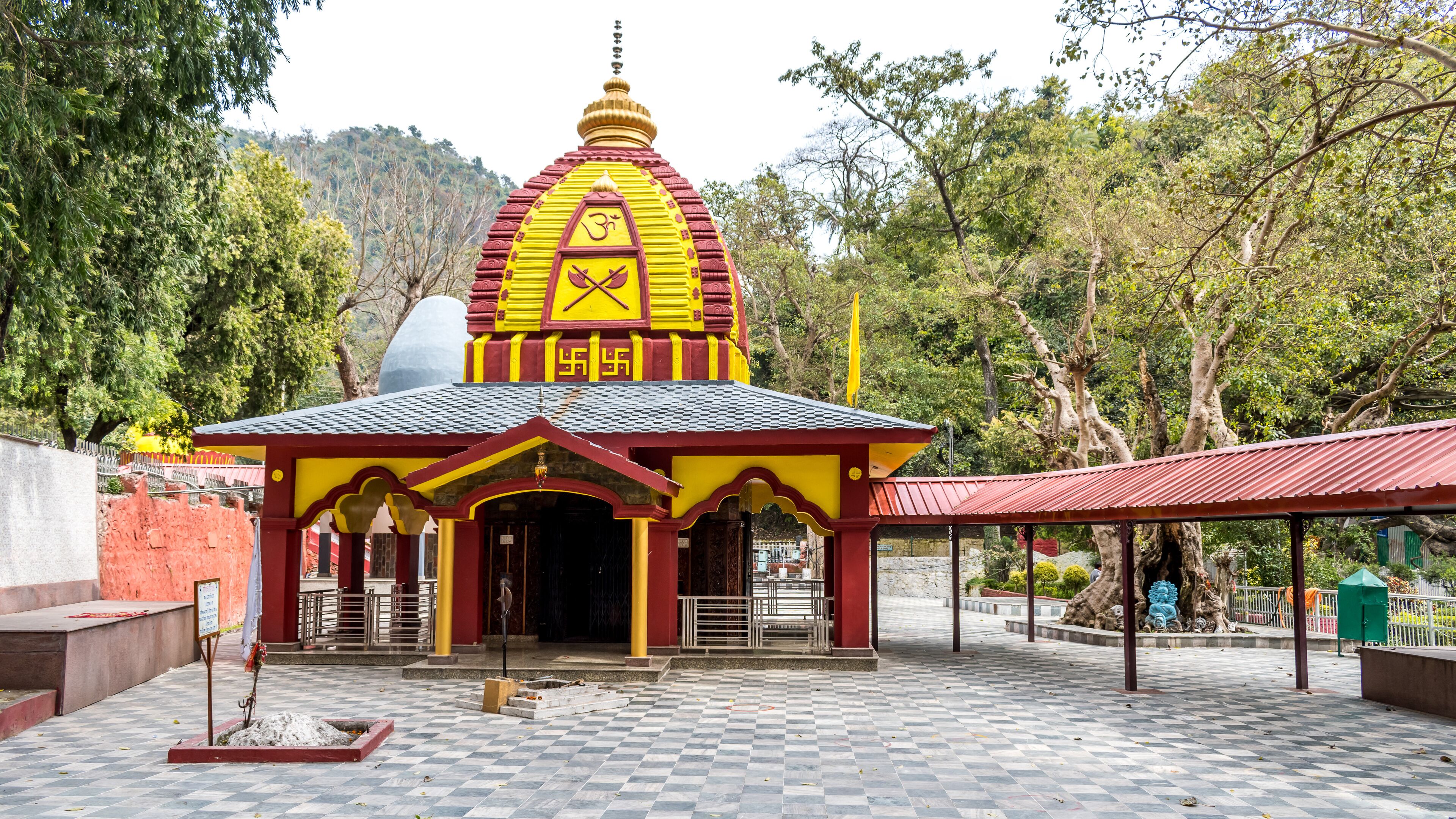A temple with a yellow and red roof, surrounded by trees and a checkered pavement.