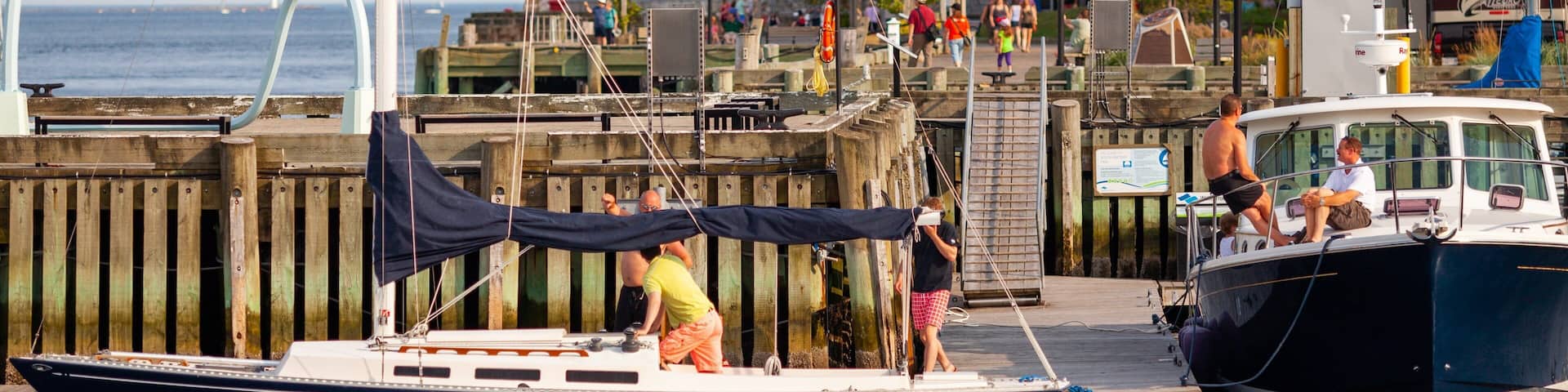A docked sailboat with people on it, a pier, and a building in the background.