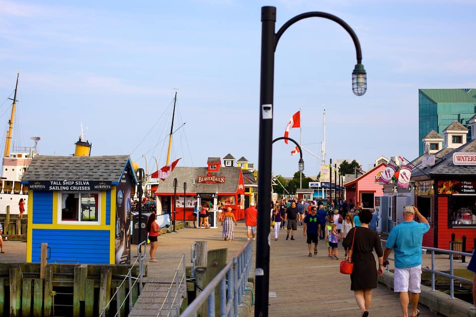 A bustling waterfront promenade with a pier, a tall ship, and various shops including a pizza restaurant.