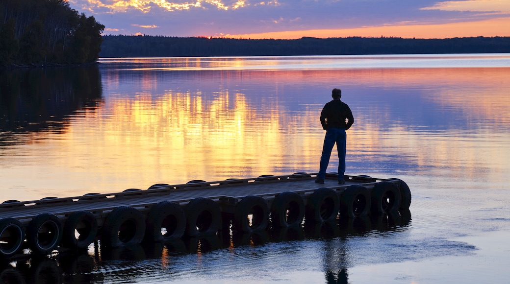 A man stands at the end of a dock watching the glowing horizon at sunset reflected in tranquil water of Gregoire Lake; Fort McMurray, Alberta, Canada