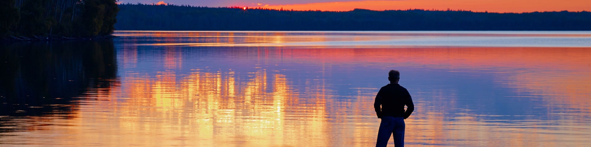 Silhouette Of A Person Standing At The End Of A Dock Looking Out Over A Tranquil Lake At Sunset, Gregoire Lake Provincial Park; Alberta, Canada