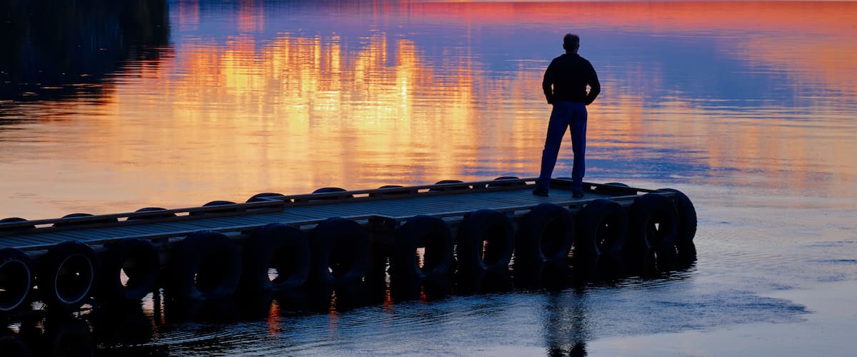 A person standing on a dock with tires, overlooking a calm lake at sunset.