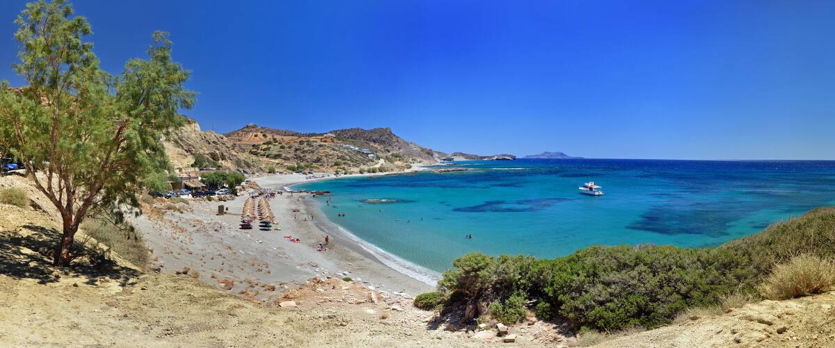 A coastal beach with clear turquoise waters, rocky cliffs, and sparse vegetation.