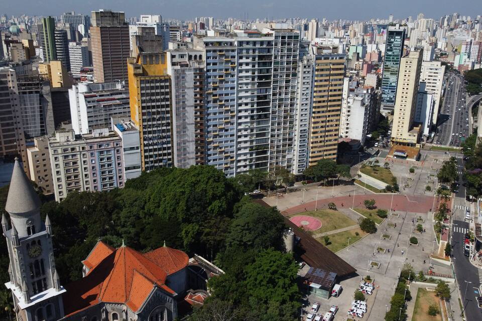 Paisagem urbana com arranha-céus, uma igreja com torre do relógio e um parque com campo esportivo.