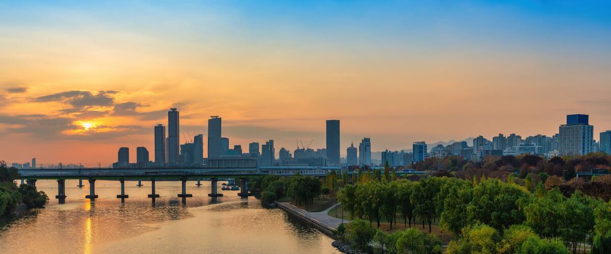A city skyline at sunset with a river and bridge in the foreground.