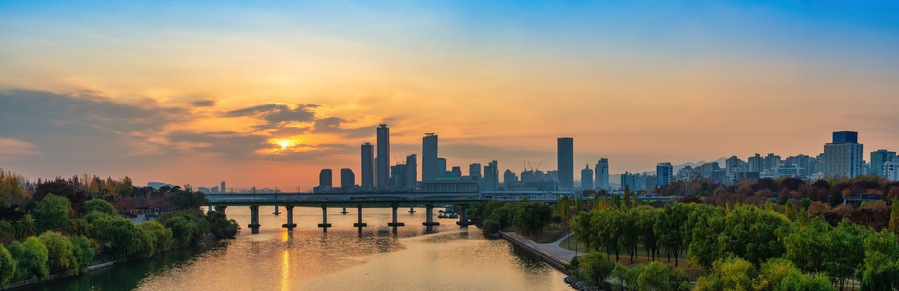 A city skyline at sunset with a river and bridge in the foreground.