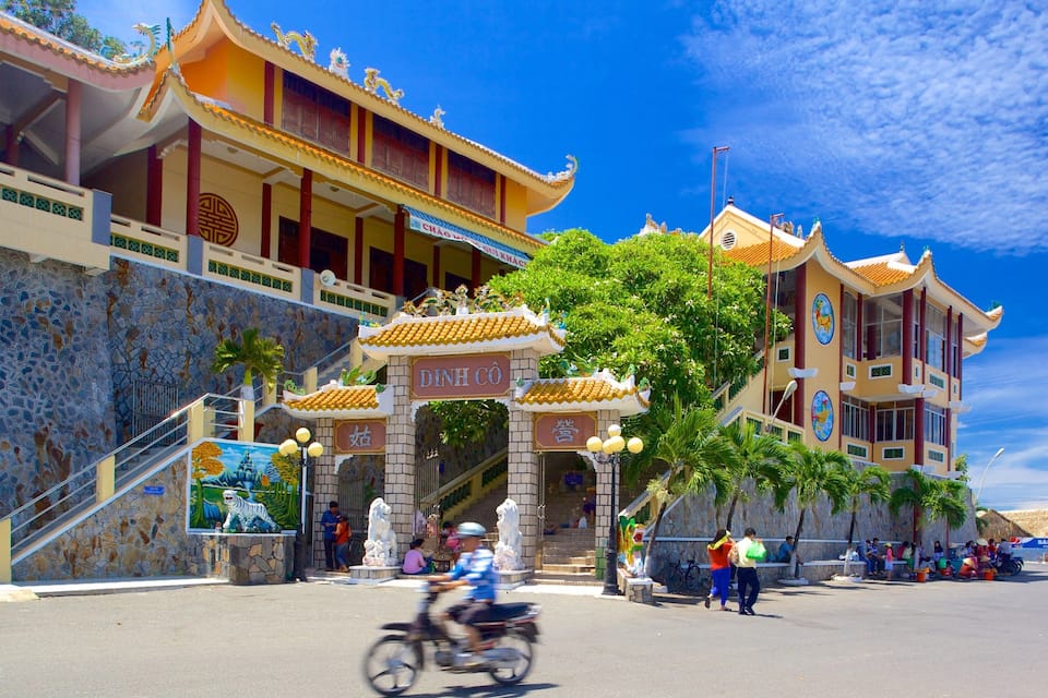 A traditional Chinese building with ornate architecture, a stone wall with mosaic tiles, and a street with pedestrians and a motorbike.