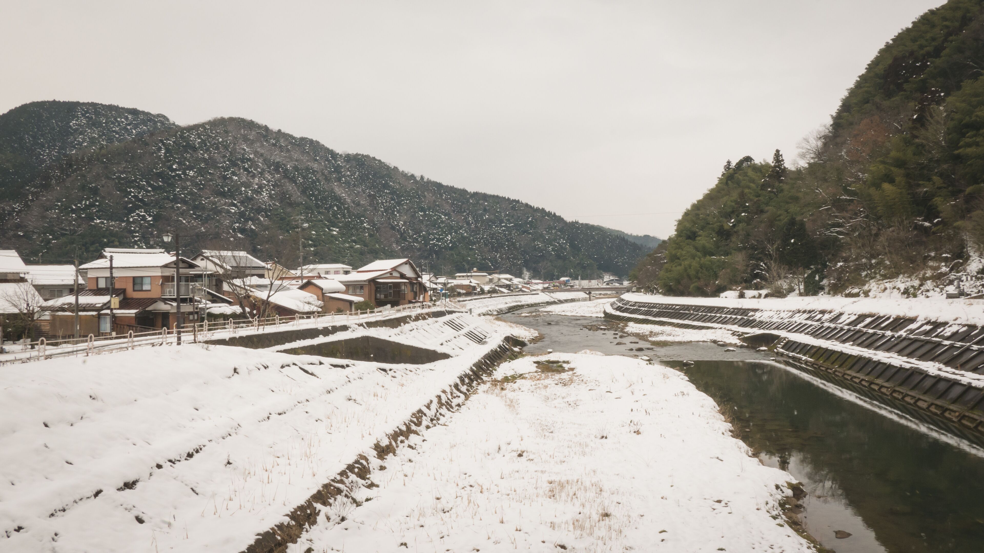 A snow-covered landscape with a river, buildings, and mountains.