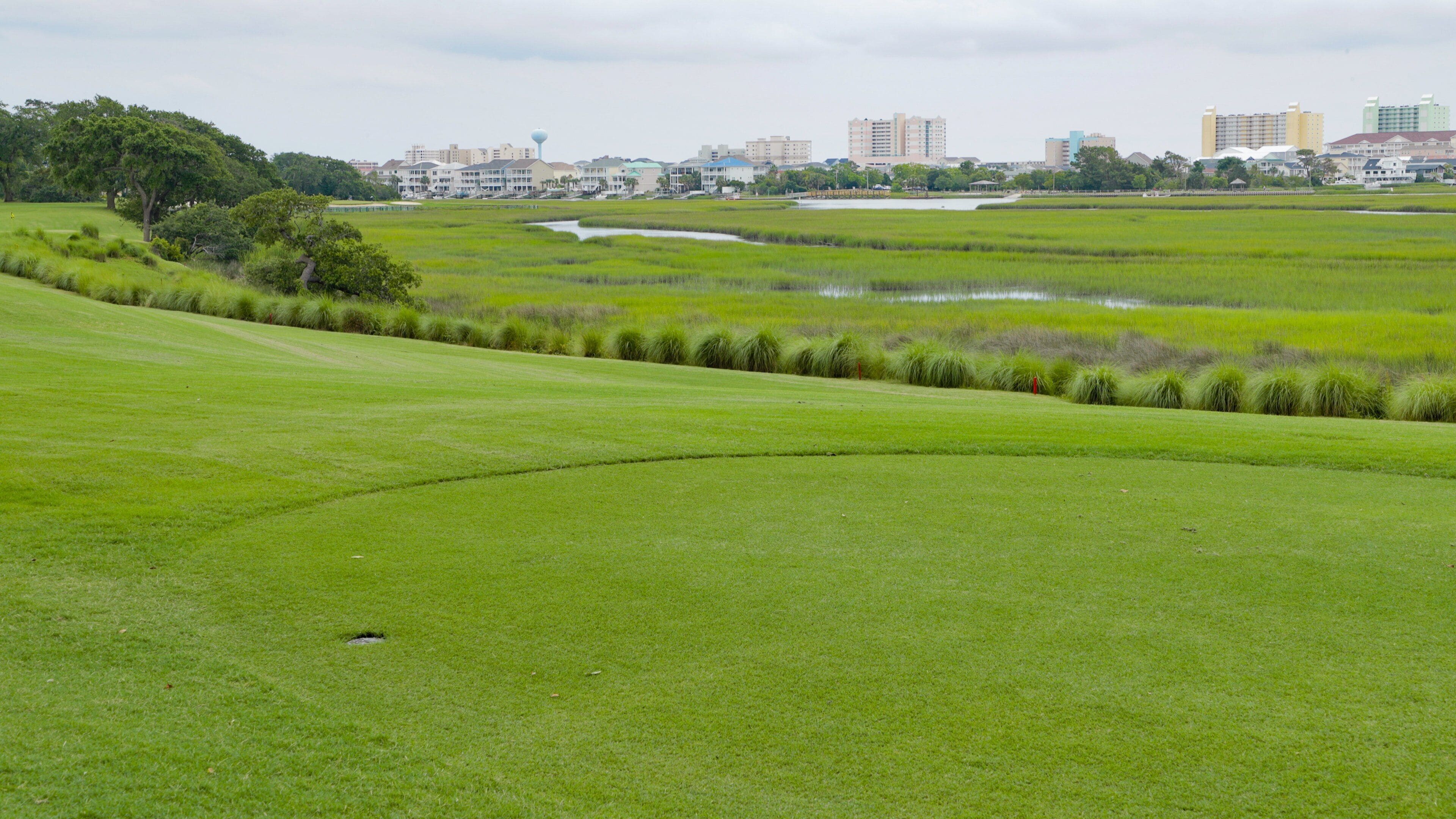 Un magnifique terrain de golf verdoyant, avec un plan d’eau et des bâtiments de la ville qui se dessinent à l’horizon.