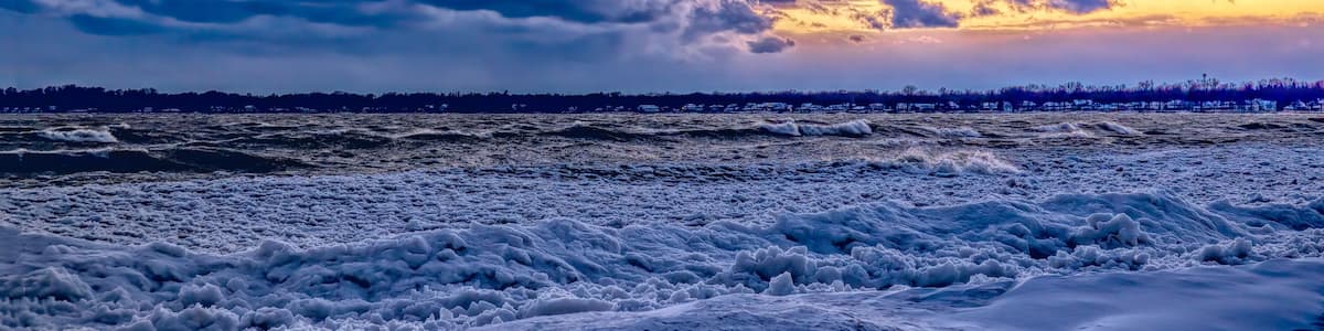 Un paysage côtier avec des vagues, un coucher de soleil et les contours d’une ville lointaine à l’horizon.