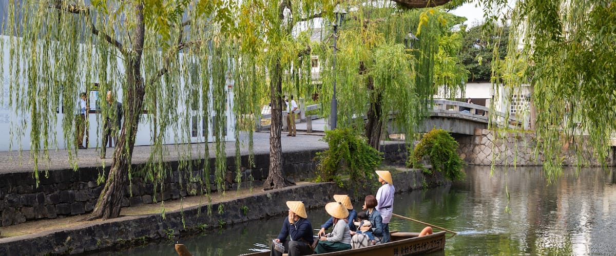 A traditional boat with passengers wearing conical hats on a calm river, surrounded by willow trees and stone walls.