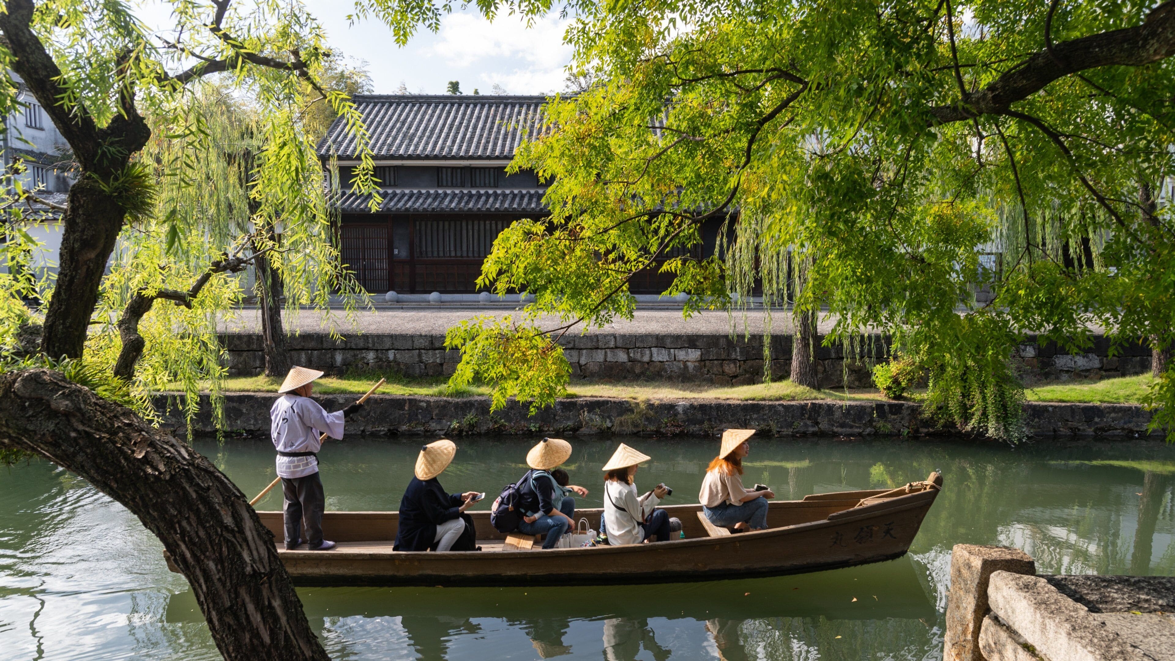 A traditional wooden boat on a calm river, surrounded by lush greenery and a historic building in the background.