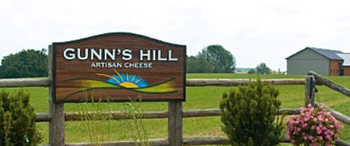 A wooden sign with 'Gunn's Hill Artisan Cheese' written on it, surrounded by a wooden fence and greenery.