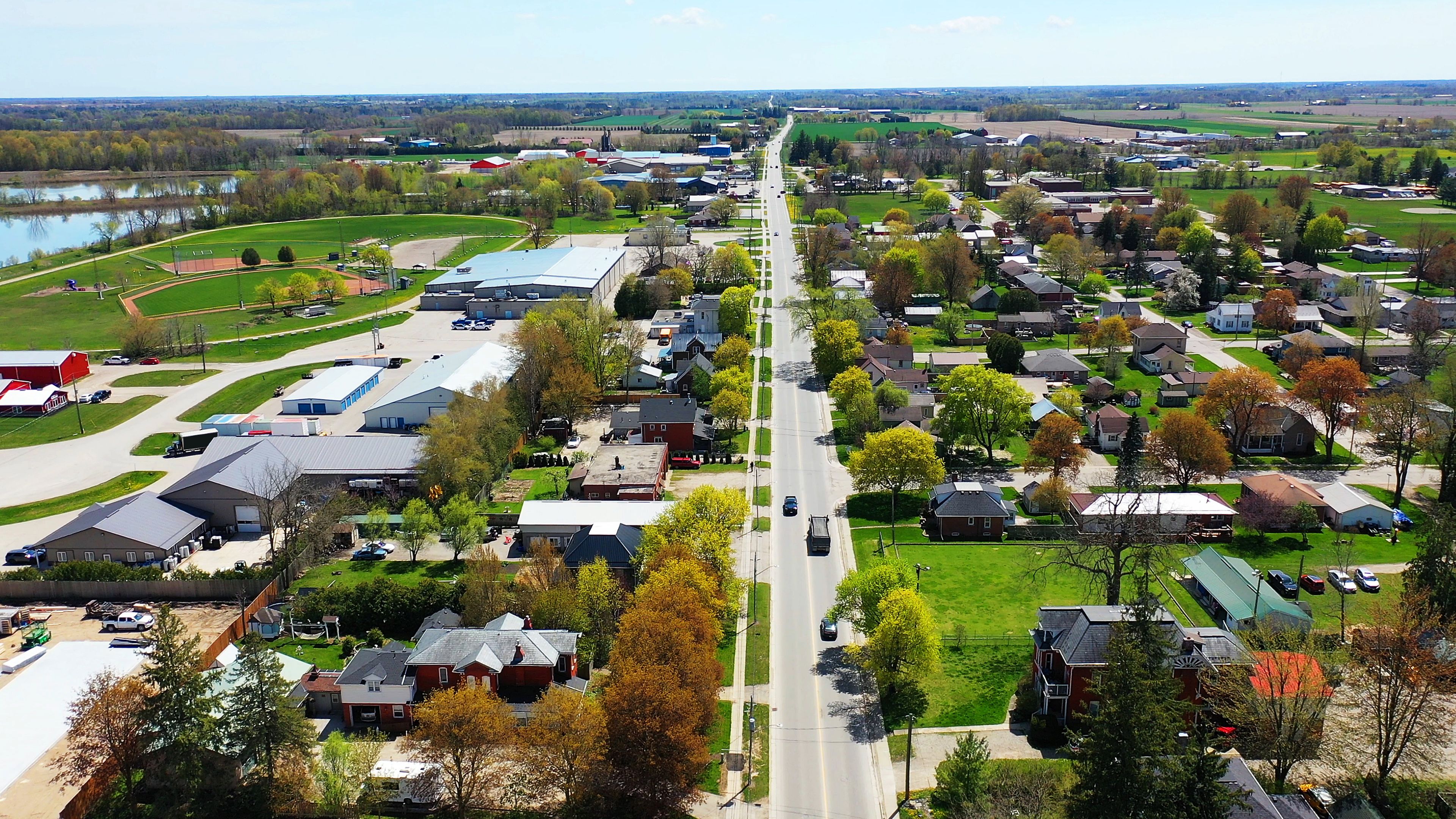 A suburban area with a main road, residential houses, a large green field, and a body of water in the background.