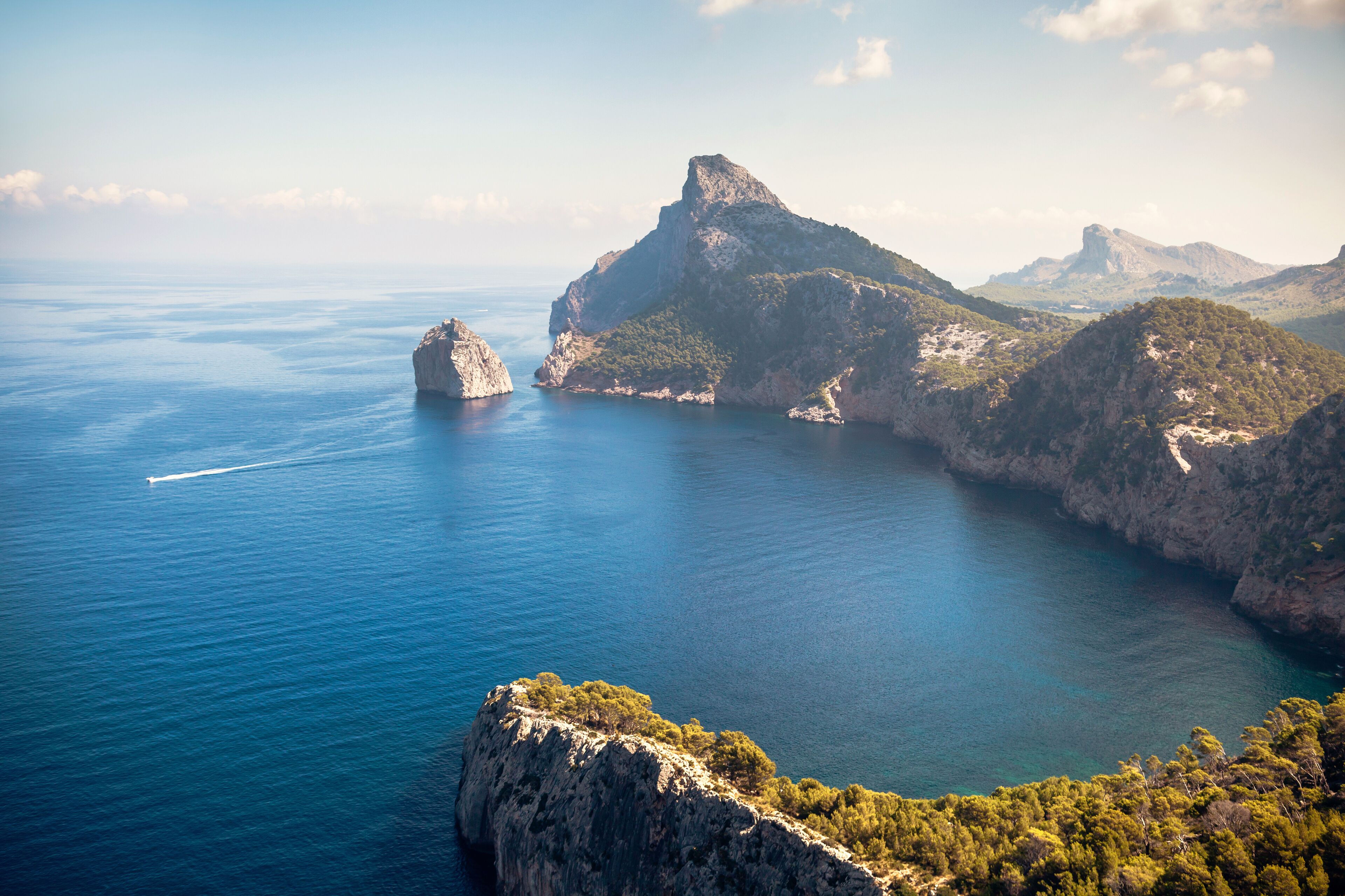 A coastal landscape with rocky cliffs, a clear blue sea, and a distant mountain peak.