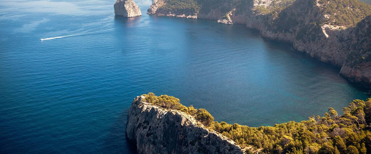 A coastal landscape with rocky cliffs, a clear blue sea, and a distant mountain peak.