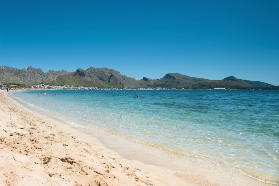 A beach with clear turquoise water, sandy shore, and rocky mountains in the background.