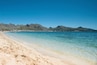 A beach with clear turquoise water, sandy shore, and rocky mountains in the background.