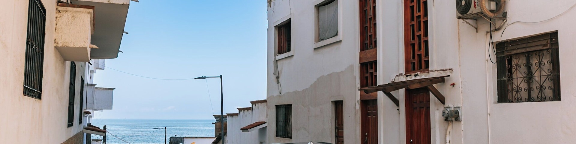 A narrow street with parked cars, a building with a white facade, and a view of the sea in the background.