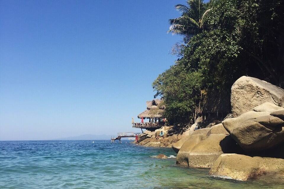 A clear blue sea, rocky shore, and a thatched-roof structure on a cliff.