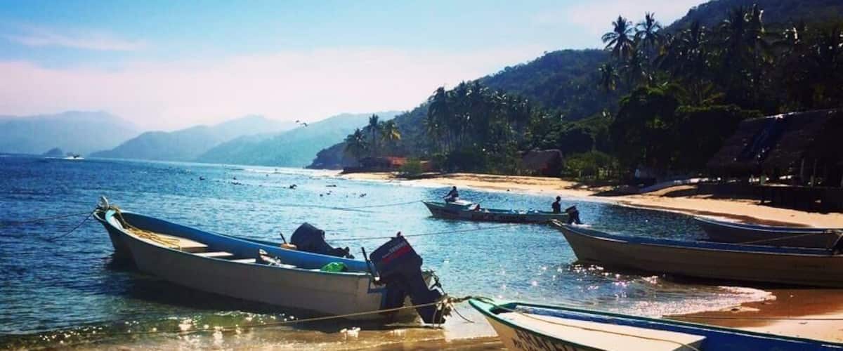 Une plage où plusieurs bateaux sont amarrés près du rivage, entourée de palmiers et avec des montagnes en arrière-plan.