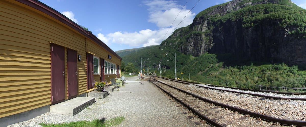 A train station with a yellow building, gravel platform, and tracks leading into a mountainous area.