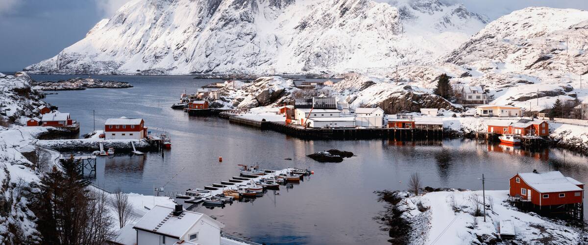 A snowy coastal village with houses, boats, and mountains in the background.