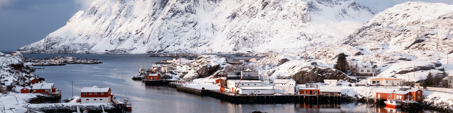 A snowy coastal village with houses, boats, and mountains in the background.