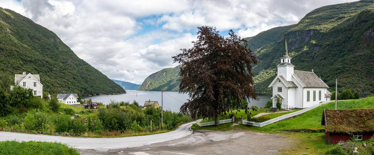 A white church with a steeple, surrounded by lush green hills and a lake.