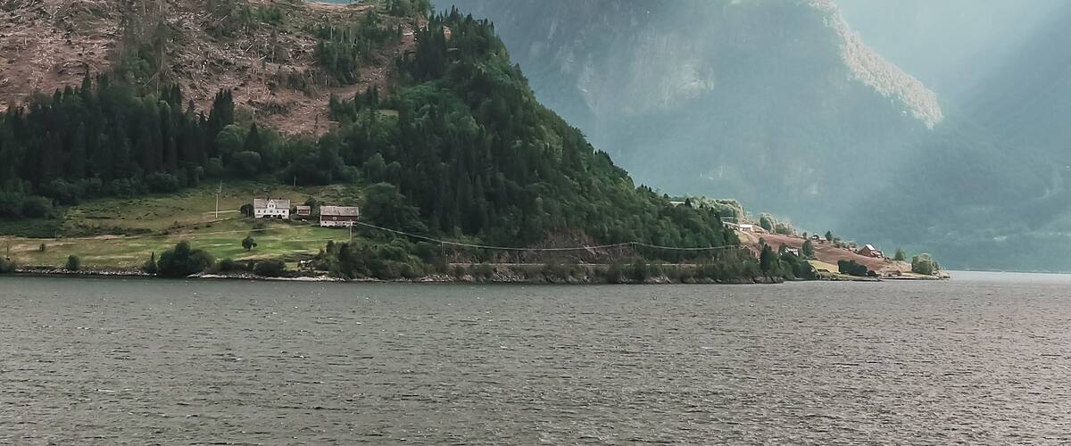 A lake surrounded by mountains with patches of snow, a small settlement with buildings, and a cloudy sky.