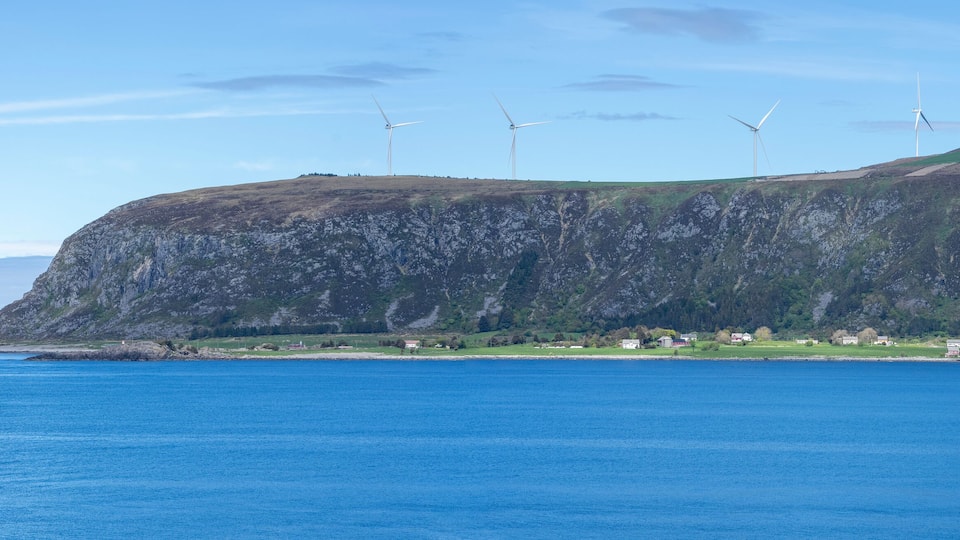 A coastal landscape with a steep cliff, a small settlement, and multiple wind turbines.